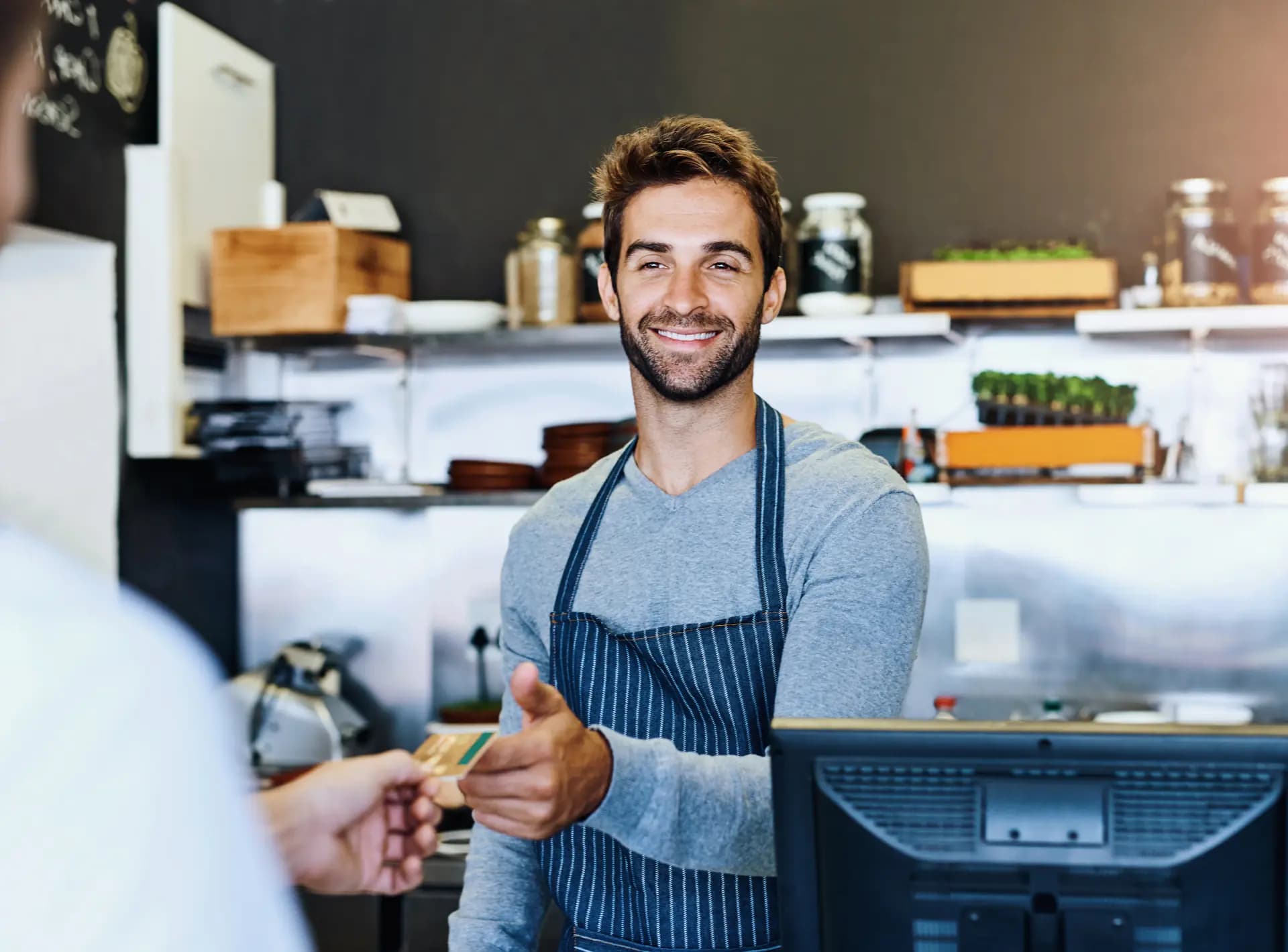 Barista sorridente recebendo cartão de crédito de cliente em balcão de cafeteria, com prateleiras e utensílios ao fundo